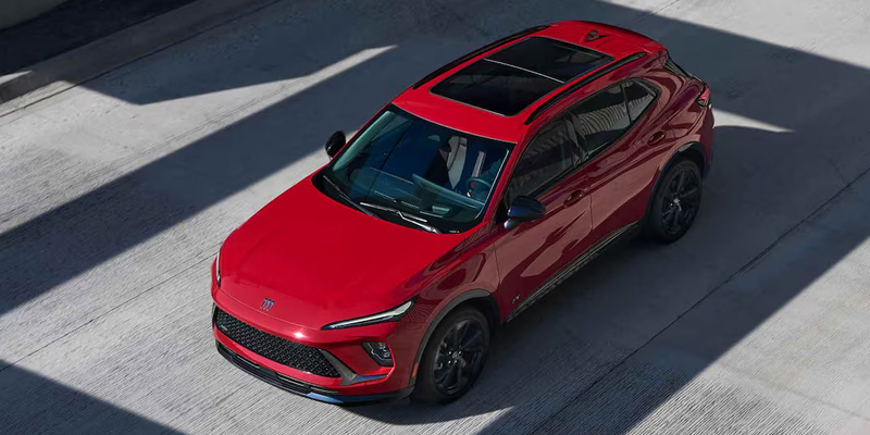 A deep red 2026 Buick Envision SUV has a sunroof and is parked on the salesroom floor with sunlight shining on it.