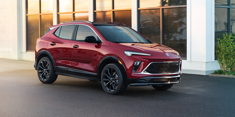 A cranberry red 2026 Buick Encore GX SUV is parked in front of a lovely white building with many large windows.
