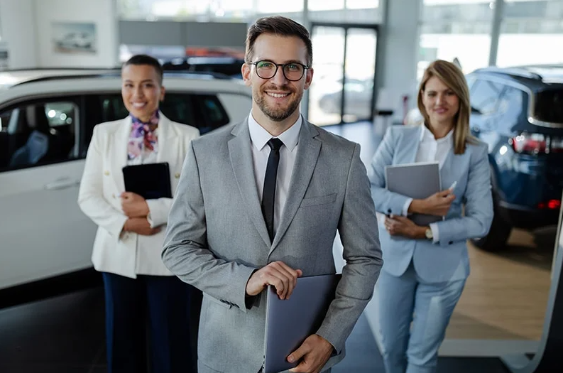 In a Buick dealer salesroom 2 lovely suited sales women and 1 suited sales man stand smiling and ready to facilitate your purchase in front of a black and a white SUV.